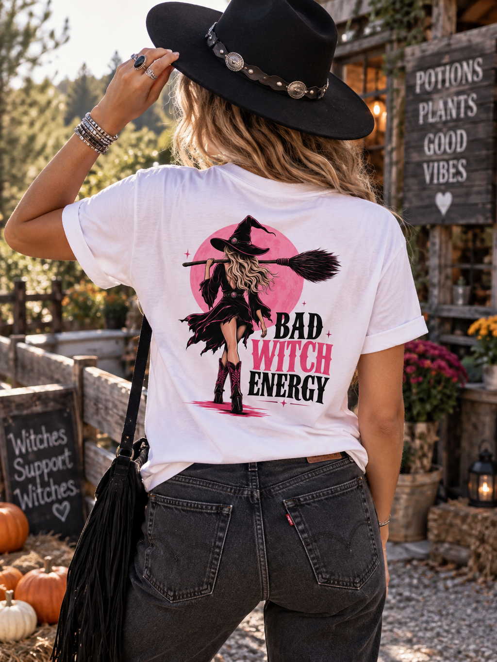 A woman wearing a white graphic t-shirt with a 'Bad Witch Energy' witch illustration on the back, paired with dark gray jeans and a black hat, photographed outdoors in a rustic autumn setting.