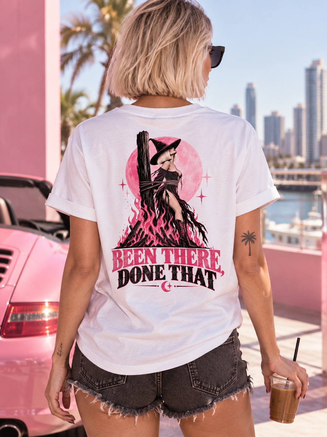 A woman viewed from behind wearing a white graphic t-shirt with a large pink and black witch-themed print that reads 'BEEN THERE, DONE THAT'. She is wearing denim shorts and holding an iced coffee on a sunny rooftop with a pink car and city skyline in the background.