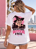A woman wearing a white graphic t-shirt with a 'Resting Witch Face' print, photographed from behind in an outdoor coastal setting with a pink building and ocean in the background.